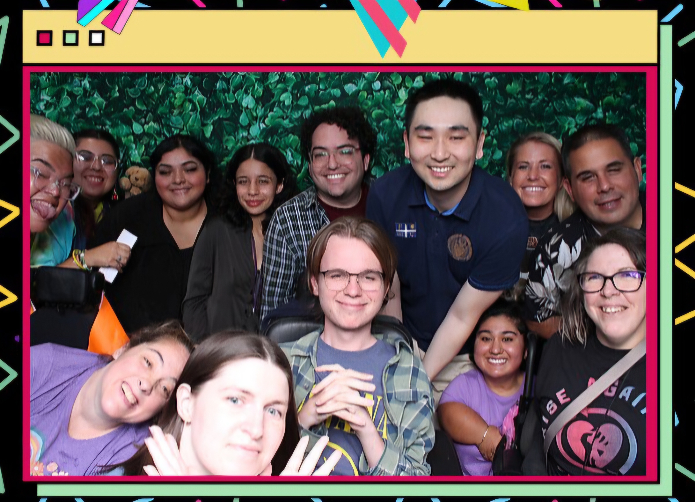 A cheerful group of people pose closely together in front of a leafy green backdrop, smiling and enjoying the moment.